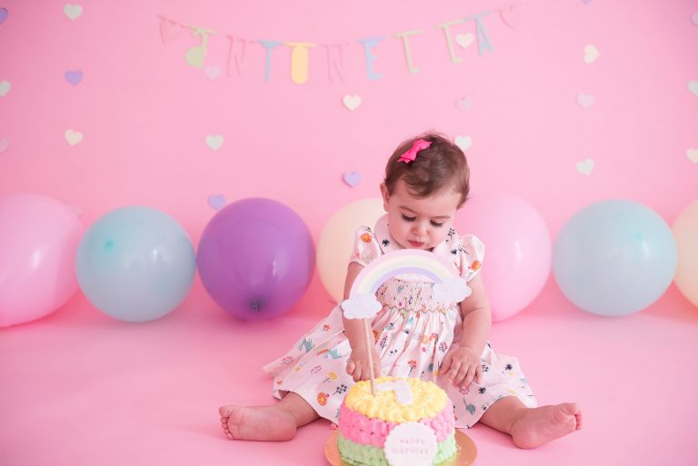 bolo colorido com arco-íris bebê sentada em cadeira com fundo rosa com balões candy colors smash the cake laura alzueta estúdio de fotografia são paulo ensaio de fotos de aniversário de 1 ano de bebê menina
