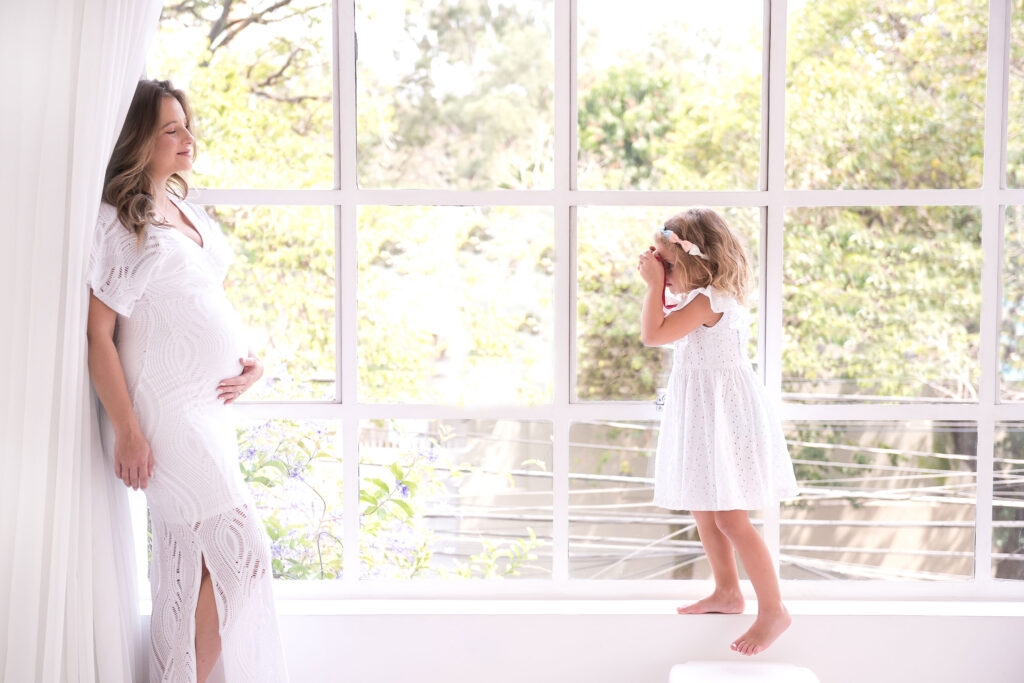 Uma mulher de vestido branco, grávida, próxima da janela, e uma criança de vestido branco olhando para ela.